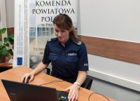 Na zdjęciu widoczna umundurowana policjantka, która siedzi przy biurku i prowadzi lekcje zdalne za pomocą laptopa. W tle baner z napisem Komenda Powiatowa Policji w Prudniku.