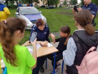 policjantka siedzi na krześle przy stole, obok niej siedzi dziewczyna i rozmawiają. Przed nimi dwie dziewczynki odwrócone plecami, obok stoi policjant i robi zdjęcie telefonem